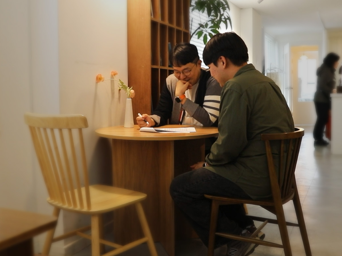 보험 계약, 손해보험 설계사, 수익 창출, 보장 분석, 간단한 부업, 따뜻한 우드 톤의 원형 테이블에 앉아 안경을 쓴 멘토가 카키색 셔츠를 입은 파트너에게 펜으로 서류를 짚어가며 1:1로 친절하게 상담을 진행하고 있는 모습