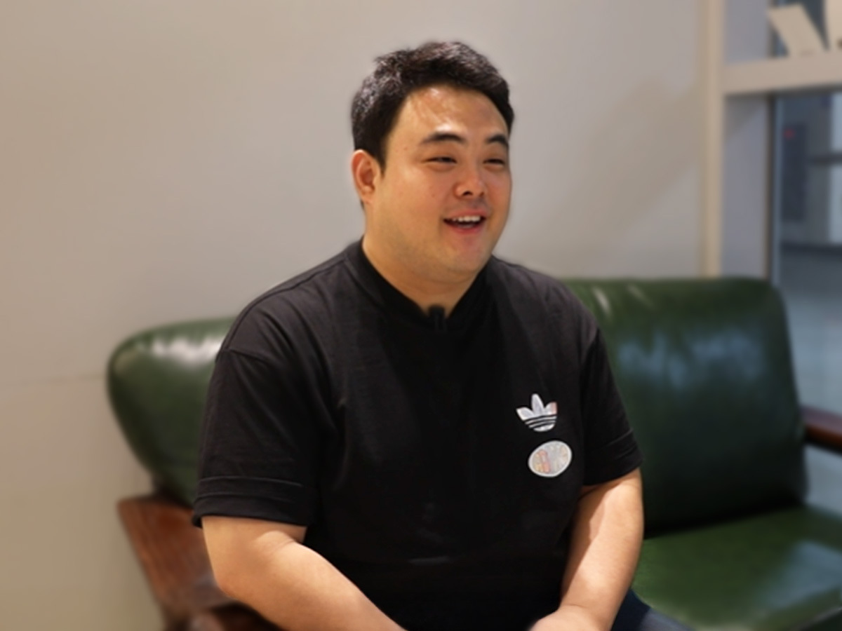 검은색 반팔 티셔츠를 입은 남성이 초록색 가죽 소파에 앉아 밝게 미소 지으며 인터뷰에 응하고 있는 모습