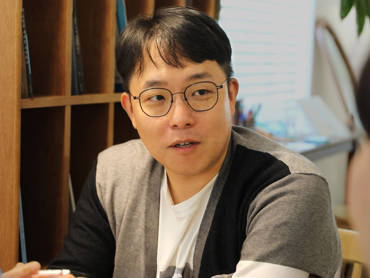 책장이 보이는 실내에서 안경을 쓴 남성이 미소를 지으며 상대방과 대화하고 있는 모습을 담은 인터뷰 스틸 컷