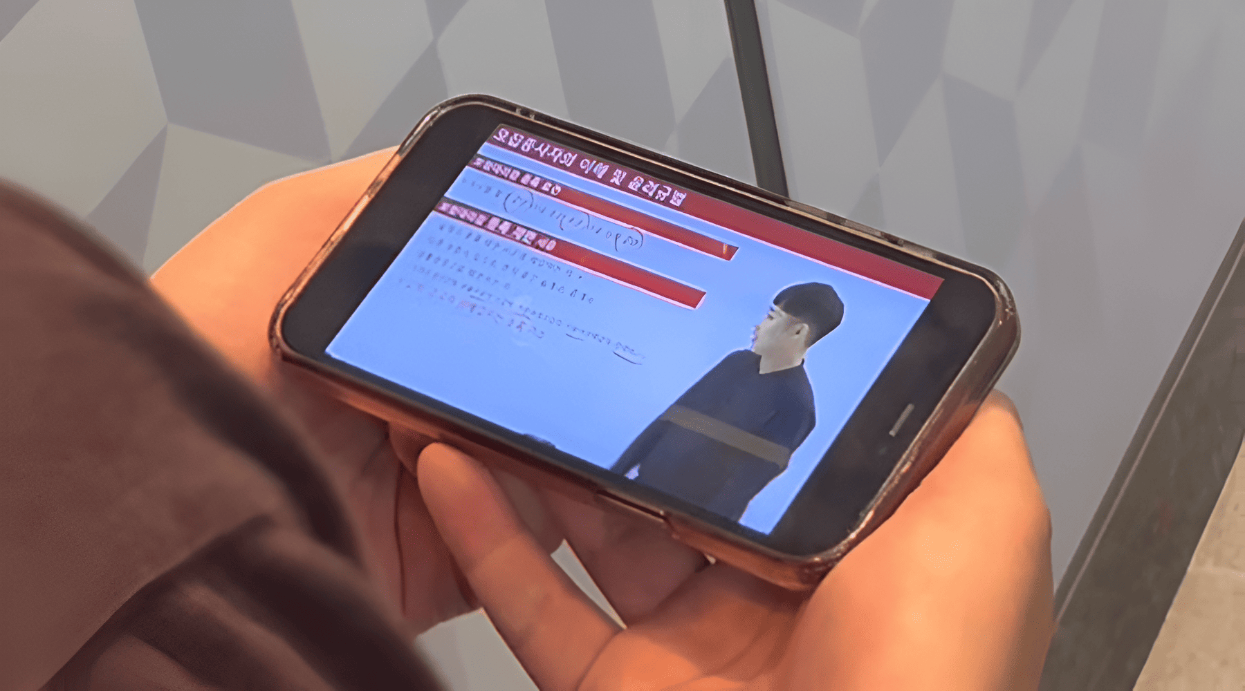 보험설계사 자격증, 기하학적 벽면 배경 앞에서 스마트폰을 양손으로 들고 메리츠 파트너스 '모집종사자의 이해 및 윤리규범' 동영상 강의를 수강하고 있는 직장인 학습자의 사진